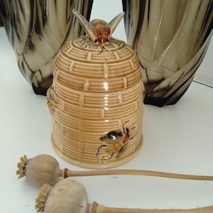 May include: A ceramic honey pot shaped like a beehive with a lid featuring a sculpted bee, accompanied by two dark translucent glass vases with a twisted design and three dried poppy seed pods with long stems arranged in the foreground.