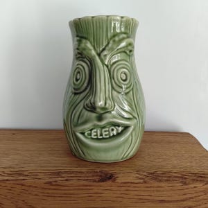 Può includere: Un vaso in ceramica verde chiaro a forma di gambo di sedano con un volto scolpito. La superficie presenta striature verticali, un naso prominente, occhi a spirale e una bocca larga con la parola CELERY incisa sul labbro inferiore.