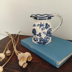 May include: A white ceramic pitcher with a hexagonal base and a scalloped rim, featuring a blue floral pattern. The pitcher has a curved handle and a pouring spout, with intricate blue botanical illustrations decorating the exterior surface.