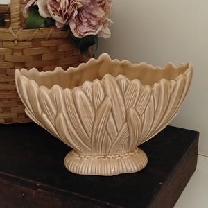 Könnte beinhalten: Eine beige Keramikschale mit einer strukturierten Außenseite aus überlappenden, blütenblattartigen Formen und einem gewellten Rand. Die Schale hat einen ausgestellten Körper, der sich zu einem abgerundeten, gerippten Sockel verjüngt.
