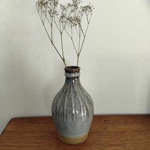 May include: A ceramic vase with a bulbous body and a narrow neck, featuring a vertical ribbed texture. The glaze is a mottled blend of grey, white, and brown tones, with a solid tan base. Dried white baby's breath flowers are placed inside the vase.