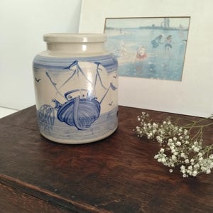 May include: A cylindrical ceramic jar with a light beige base and blue hand-painted illustrations of a sailboat on the water, fishing nets, and birds. The jar features a smooth, rounded rim and a wide opening, showcasing a nautical theme.