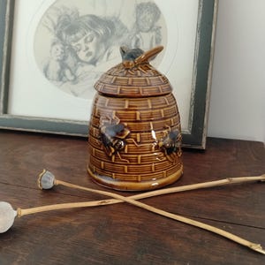 May include: A brown ceramic honey pot shaped like a traditional beehive, featuring a textured basket-weave pattern and raised bee figures on the exterior. The lid is topped with a sculpted bee, and the entire piece has a glossy finish.