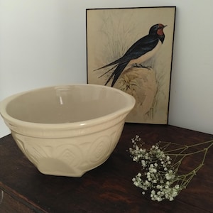 May include: A cream-coloured ceramic mixing bowl with a wide rim and a repeating embossed geometric pattern around the exterior. The bowl features a smooth, glossy finish and a deep, rounded basin, designed for kitchen use and food preparation.
