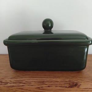 May include: A rectangular dark forest green ceramic butter dish with a smooth, glossy finish. The container features a matching lid topped with a small, centered spherical knob handle, providing a clean and functional design for kitchen storage.