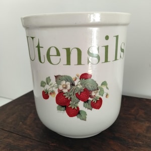 Könnte beinhalten: Ein weißer Utensilienhalter aus Keramik mit dem Wort Utensils in grüner Schrift. Die Vorderseite des Behälters ist mit einer botanischen Illustration von roten Erdbeeren, grünen Blättern und kleinen weißen Blüten verziert.