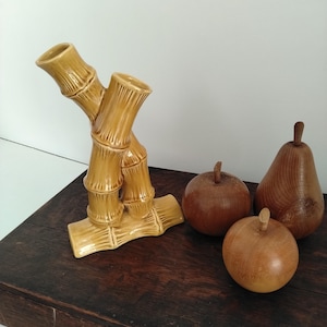 Könnte beinhalten: Eine glänzende, senfgelbe Keramikvase in Form von Bambusstangen, begleitet von drei glatten, geschnitzten Holzfrüchten, bestehend aus zwei runden Äpfeln und einer Birne, die alle sichtbare natürliche Holzmaserungen aufweisen.