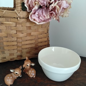 May include: A woven wooden basket containing large, dusty pink artificial peony flowers, positioned next to two small, brown ceramic deer figurines with white spots and a plain, off-white ceramic mixing bowl with a smooth, rounded rim.