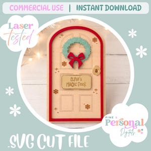 Op de afbeelding: Een boogvormige deur van licht hout met een rood frame, een blauwgroene krans met een rode strik en een rechthoekig plaatje met de inscriptie "Elfie's Magic Door". De deur is versierd met gegraveerde sneeuwvlokken, sterren en een gouden knop.