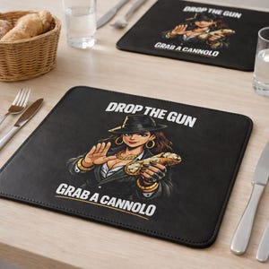 Può includere: Una tovaglietta rettangolare nera con un'illustrazione grafica di una persona con fedora e completo che tiene in mano un cannolo. Il testo "DROP THE GUN" appare sopra la figura, e "GRAB A CANNOLO" è stampato sotto in grassetto maiuscolo bianco.