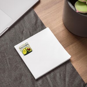 Può includere: Un blocco note quadrato bianco con una piccola grafica nell'angolo. La grafica raffigura due metà di avocado con polpa verde e nocciolo marrone, circondate da ghirigori decorativi e dal testo nero "Timing is Everything" scritto sopra.