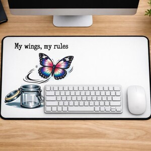 Può includere: Un tappetino da scrivania rettangolare con superficie bianca che presenta una farfalla colorata con ali rosa, viola e blu, un barattolo di vetro con coperchio e il testo nero "My wings, my rules" stampato in carattere serif in alto a sinistra.