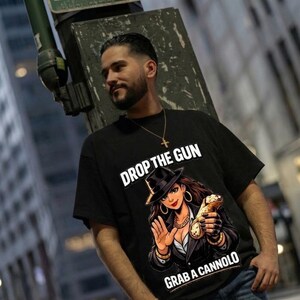 Può includere: Una t-shirt nera a maniche corte con un'illustrazione grafica di una persona che indossa un fedora e un abito, mentre tiene in mano un cannolo. Il testo "DROP THE GUN" è stampato in bianco sopra e "GRAB A CANNOLO" è stampato in bianco sotto.