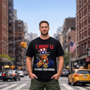 Può includere: Una t-shirt nera a maniche corte con una grafica dello Zio Sam che indossa un cilindro a stelle e strisce, una giacca blu e una collana con il simbolo del dollaro dorato. Il testo I WANT U TO PROVE THEM WRONG è stampato in rosso e bianco.