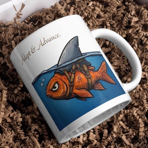 Può includere: Una tazza in ceramica bianca con un'illustrazione grafica di un pesce rosso arancione che indossa un'imbracatura con pinna di squalo. Il testo "Adapt & Advance." è stampato in nero sopra il pesce che nuota in acqua blu.