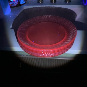 May include: A stack of three translucent red glass plates featuring a textured, ribbed pattern along the outer rim. The plates exhibit a vibrant, glowing appearance under ultraviolet light, highlighting the circular design and the glass material.