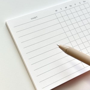 May include: A white paper habit tracker notepad with black printed lines and a grid. The top left corner features the word HABIT, while the top right displays the letters M, T, W, Th, F, S, Su. A beige pen rests diagonally across the notepad.