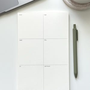 May include: A white weekly planner notepad with a grid layout featuring the days of the week labeled mon, tue, wed, thu, fri, and sat & sun in small black text. The notepad is positioned next to a matte olive green retractable ballpoint pen.