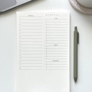 May include: A white daily planner notepad with black ink printing. The layout features a schedule column from 6 to 10, a to-do list section, a habits tracker, and a tomorrow section. The days of the week are listed as M T W T F S S.