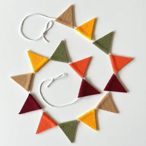 May include: A decorative bunting garland featuring a series of triangular felt flags in shades of tan, yellow, olive green, orange, and deep burgundy. The felt triangles are attached to a thin white string, which is tied into small loops at each end.