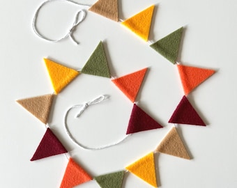 Op de afbeelding: Een decoratieve slinger met een reeks driehoekige viltvlaggetjes in de kleuren beige, geel, olijfgroen, oranje en donkerbordeaux. De vilten driehoeken zijn bevestigd aan een dun wit touwtje dat aan beide uiteinden in kleine lussen is geknoopt.