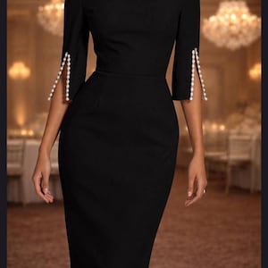 May include: A black knee-length dress featuring a boat neckline, three-quarter length sleeves, and a fitted silhouette. The sleeves have a vertical slit detail along the outer forearm, which is embellished with a row of small white pearls.