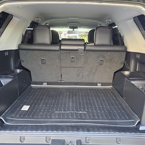 May include: The interior cargo area of a vehicle featuring a black rubber floor mat with a textured surface and raised edges. Two black side storage panels are installed along the interior walls, positioned in front of the rear passenger seats.