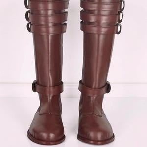 May include: A pair of knee-high dark brown boots featuring a smooth finish, rounded toes, and a series of three horizontal straps with buckles around the upper calf area, plus a single strap with a buckle positioned around the lower ankle section.