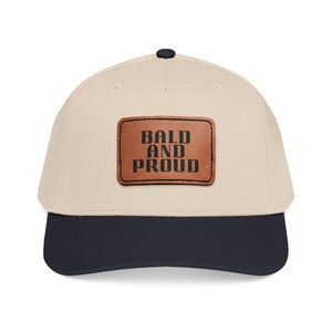 May include: A baseball cap with a cream-colored crown and a navy blue brim. A brown leather patch on the front displays the words "BALD AND PROUD" in black block letters. The cap has a black button on top.