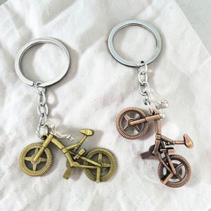 May include: Two metal bicycle keychains with silver-toned split rings and chains. One keychain features a brass-toned bicycle charm, while the other features a copper-toned bicycle charm. Both charms display detailed frames, handlebars, and wheels.