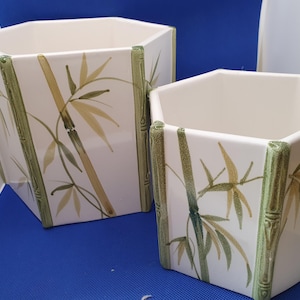 May include: Two hexagonal ceramic planters with a glossy white finish, featuring painted green and tan bamboo stalks and leaves. The edges of each planter are moulded to resemble textured green bamboo segments, creating a cohesive design.