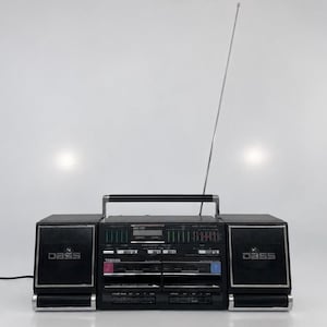 May include: A black Toshiba portable stereo system featuring dual cassette decks, a radio tuner, and two detachable speakers with DBSS branding. The unit includes a top-mounted carrying handle and a long, thin, silver-coloured telescopic antenna.