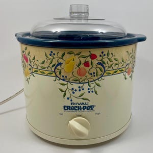 May include: A cream-coloured Rival Crock-Pot stoneware slow cooker with a clear plastic lid. The exterior features a decorative border of painted fruit and vines. A single white control knob is positioned at the bottom with Off, Low, and High settings.