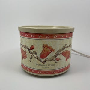Może przedstawiać: Cylindryczny podgrzewacz Rival Potpourri Crock z beżową krawędzią i podstawą. Korpus ma kremowe tło z powtarzającym się kwiatowym wzorem pomarańczowych i czerwonych tulipanów na zielonych łodygach. Napis Potpourri Crock RIVAL jest wydrukowany na brązowo.