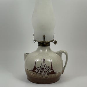 Könnte beinhalten: Eine Öllampe aus Keramik mit einem runden, gesprenkelten beigen Sockel, der eine braune Hausillustration und einen Henkel aufweist. Die Lampe hat einen Messingbrenner und einen hohen, mattierten Glaszylinder, der oben in Klarglas übergeht.
