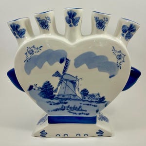 May include: A white ceramic tulip vase shaped like a heart with five individual spouts. The vase features blue hand-painted illustrations of a Dutch windmill, clouds, and floral motifs. It has a wide base and two blue side handles for stability.