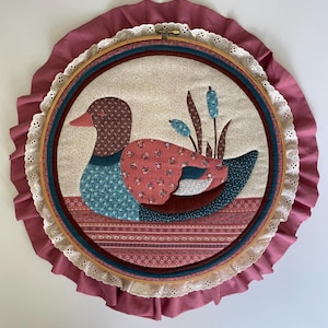 May include: A circular fabric wall hanging featuring a quilted duck appliqué in shades of brown, blue, and pink floral patterns. The piece is mounted in a wooden embroidery hoop, framed by a white eyelet lace trim and a ruffled dusty rose border.