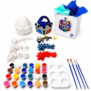May include: A craft kit featuring unpainted and painted dragon figurines, two egg-shaped containers, a white box labeled 3D COLOR LAB with blue and teal tissue paper, a grid of twenty-four small paint pots, a white paint palette, and four paintbrushes.