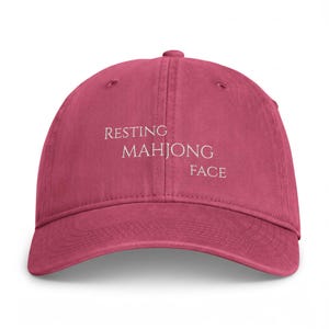 May include: A dark pink baseball cap with a curved brim and a six-panel construction. The front of the cap features the white embroidered text "RESTING MAHJONG FACE" arranged in three staggered lines across the center of the crown.