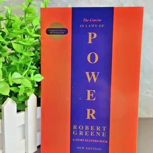 Op de afbeelding: Een hardcover boek getiteld "The Concise 48 Laws of Power" van Robert Greene. Het boek heeft een oranje omslag met een verticale blauwe streep in het midden. De tekst "POWER" staat in grote gele letters. Een "International Bestseller" sticker staat op de omslag.