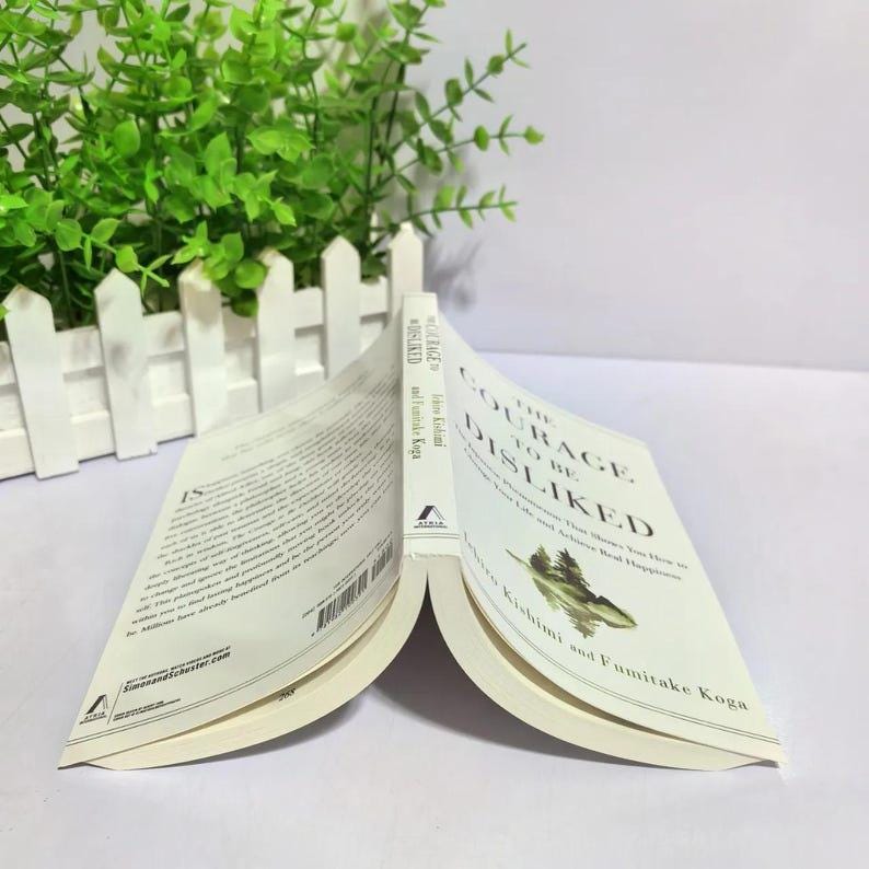 Peut inclure: Livre ouvert intitul&eacute; "Le Courage d'&ecirc;tre d&eacute;test&eacute;" par Ichiro Kishimi et Fumitake Koga. Le livre a une couverture blanche avec du texte noir et une illustration verte. Les pages sont de couleur cr&egrave;me.
