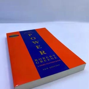 Op de afbeelding: Een boek getiteld "POWER" van Robert Greene. De omslag is oranje met een blauwe middenstreep. De titel en de naam van de auteur zijn in het wit. De woorden "NEW EDITION" zijn ook zichtbaar.
