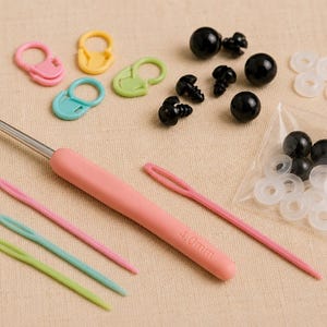 May include: A selection of crochet and amigurumi supplies. The image shows a pink handled 4.0mm crochet hook, yarn needles, stitch markers, black safety eyes, and white washers. The tools are in various colours, including pink, green, and blue.