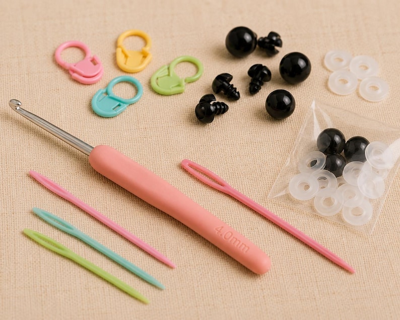 May include: A selection of crochet and amigurumi supplies. The image shows a pink handled 4.0mm crochet hook, yarn needles, stitch markers, black safety eyes, and white washers. The tools are in various colours, including pink, green, and blue.