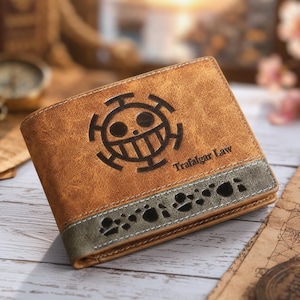 Puede incluir: Una cartera plegable de color tostado con textura, que presenta un emblema de calavera y tibias cruzadas en marrón oscuro, el texto "Trafalgar Law" y una banda horizontal gris en el borde inferior decorada con pequeñas formas oscuras irregulares.