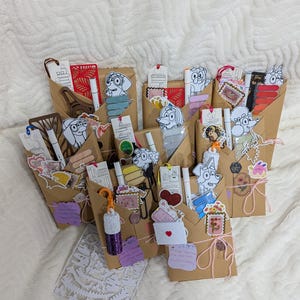 May include: A collection of brown paper gift bags, each containing a variety of stationery items including white markers, colorful stickers, die-cut paper bookmarks featuring cartoon dog characters, and small decorative tags tied with string.