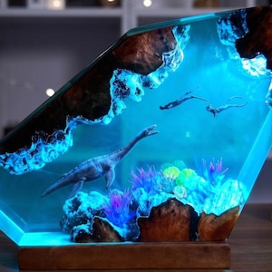 Könnte beinhalten: Eine trapezförmige Lampe aus Harz und Holz mit einer Unterwasserszene, die einen Plesiosaurus, zwei Taucher und bunte Wasserpflanzen zeigt. Das Stück wird durch ein helles blaues Licht beleuchtet, das die strukturierten Holzkanten hervorhebt.