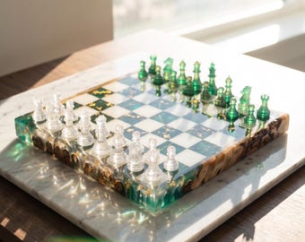 Op de afbeelding: Een vierkante schaakset met een bord van afwisselend witte en doorschijnend groene harsvierkanten met goudvlokken. Het bord heeft een rand van natuurlijk hout. Eén set schaakstukken is helder, de andere set is doorschijnend groen.