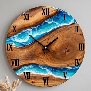 May include: A round wall clock featuring a natural wood grain slab with two flowing blue and white resin sections resembling ocean waves. The clock face displays black Roman numerals and two black hands at the center.