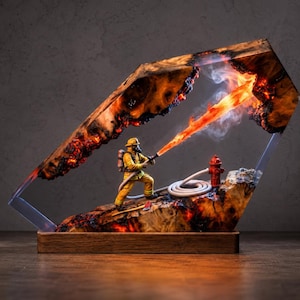 May include: A decorative lamp featuring a miniature firefighter in yellow gear spraying a stream of orange light from a hose. The scene is encased in a frame of natural wood with charred edges and translucent resin, set on a rectangular wood base.